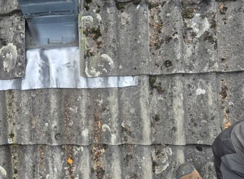 Verouderde betonnen dakpannen met zichtbare mos- en korstmosgroei rond een loodslab, als gevolg van langdurige blootstelling aan vocht en weersinvloeden.